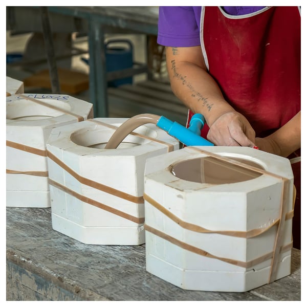 Person wraps a white ceramic item with brown tape, using blue scissors, on a workbench.