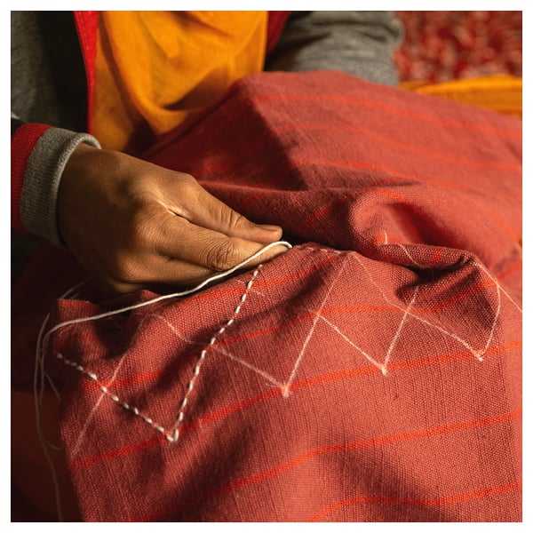 A person stitches a vibrant red, plaid-patterned cushion cover by hand. The fabric is cotton; the cover discrete stripes and a geometric design.