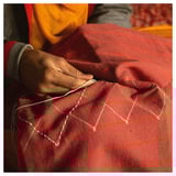 A person stitches a vibrant red, plaid-patterned cushion cover by hand. The fabric is cotton; the cover discrete stripes and a geometric design.
