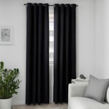 Black curtains with eyelets hang by a window, blocking light and providing privacy. A large plant and light gray sofa are nearby.