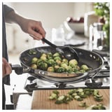 A person stirs brussels sprouts in a black MIDDAGSMAT frying pan on a gas stove. Wooden cutting board with sprouts nearby.
