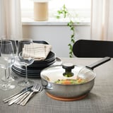 A modern kitchen table setup with a sauté pan, plates, glasses, and cutlery, ready to serve a meal. The pan has a glass lid on a trivet.