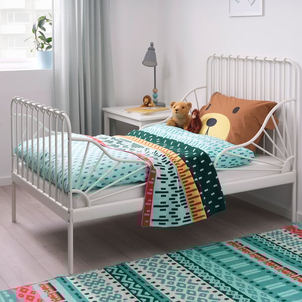 Colorful kids bedroom with white metal MINNEN bed, brown teddy bear pillow, green blankets, and a small nightstand. Bright decor, inviting and cozy.