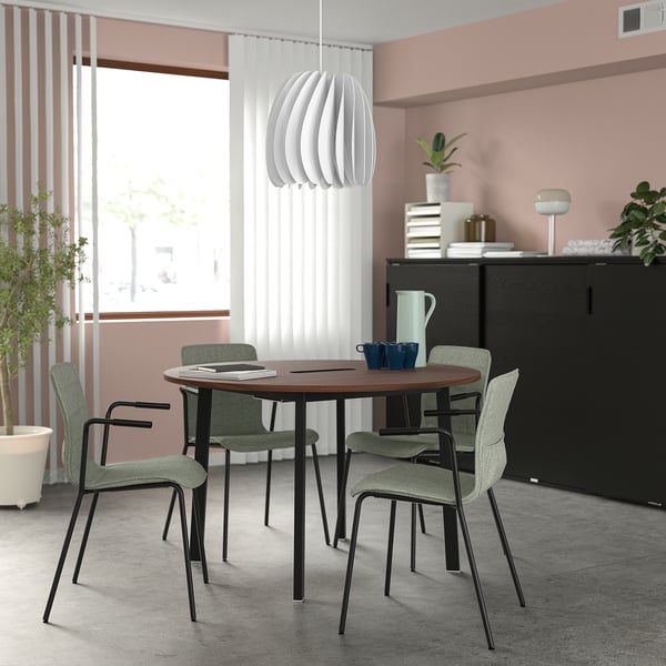 Small round MITTZON conference table with four chairs, white pendant light, and black cabinet, set against pink walls with large windows.