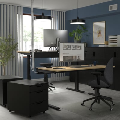Home office with MITTZON sit/stand desk, black furniture, plants, grey chair, large monitor, laptop, blue walls & big window.