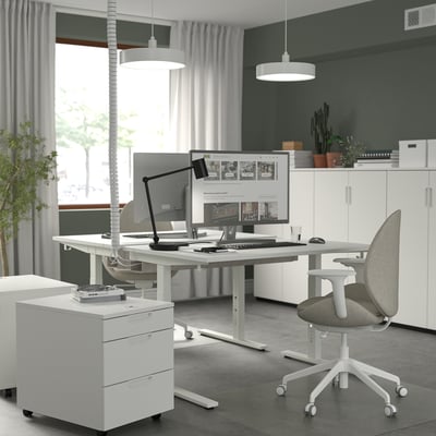 Modern home office with height-adjustable desk, dual monitors, office chair, and plants.