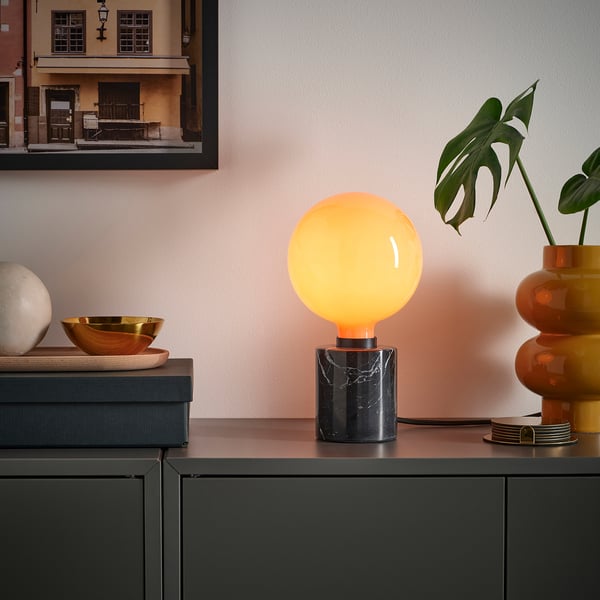 Orange MOLNART bulb in black marble base, with golden bowl, white sphere, plant, and picture.