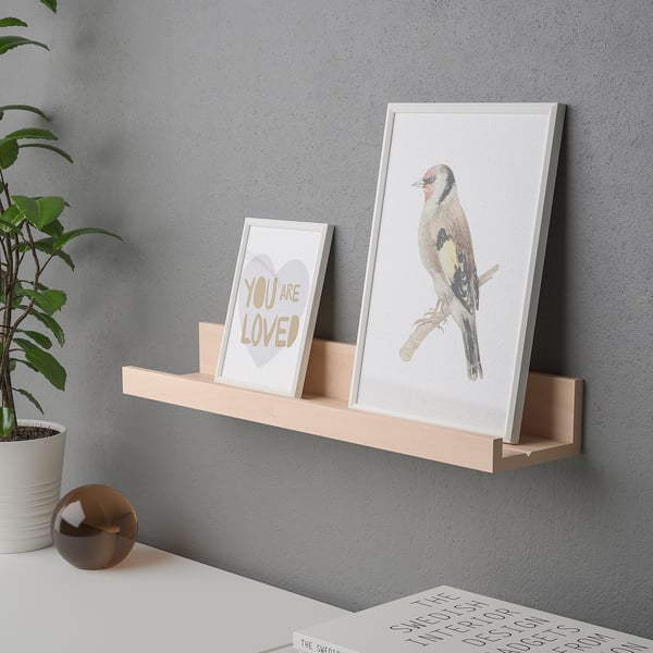 A wall shelf with a groove holds framed art. The groove supports small frames upright.