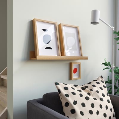 Light wood MOSSLANDA shelf holds two framed arts, one small red dot art below. Nearby, grey sofa holds spotted pillow. Modern decor.