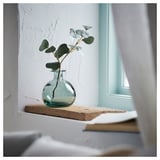 Small glass vase with green-blue tint on rustic wooden shelf. Contains single stem eucalyptus.