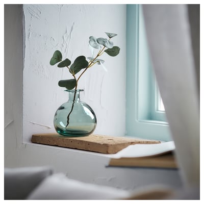 Small glass vase with green-blue tint on rustic wooden shelf. Contains single stem eucalyptus.