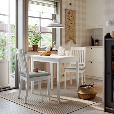 Cozy dining nook with white table, beige cushions. Baskets & teapot on table. Bright kitchen with large window.