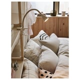 A minimalist white NÄVLINGE lamp clamped to a headboard, illuminating a bed with white and patterned pillows.