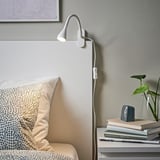 White NÄVLINGE lamp beside bed, focused light, plant, books.