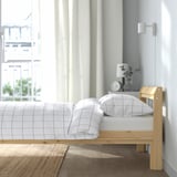 Minimalist bedroom with wooden bed, checkered linens, and bedside lamp. Natural light streams through curtains.