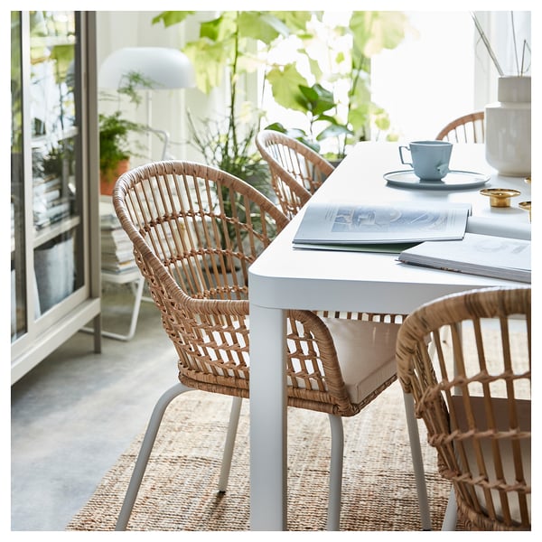 Gray wicker chair with bamboo, books, plates, and blue mug near window with plants.