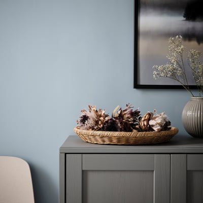 Wicker basket with dried flowers on dark cabinet, beside vase, light blue wall, framed picture.