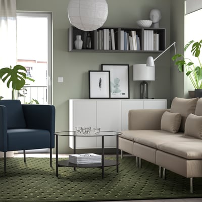 Modern living room with sage green walls, brown couch, blue chair, green rug, glass table, and plants.