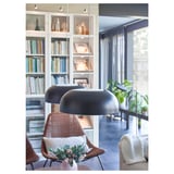 Modern living room with glass bookshelf, two stacked black NYMÅNE lamps, wicker chairs with cushions, and a window with plants.