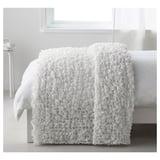 A white OFELIA bed blanket, textured and cozy, draped over a bed, suitable for 180 cm beds.