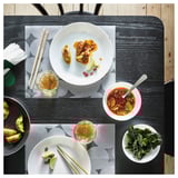 Dinner setup with white plates and bowls, chopsticks, colorful food on dark wooden table.