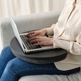 Person using a laptop on a round cushioned tray, likely the ÖJULF laptop support, while sitting on a light-colored sofa.