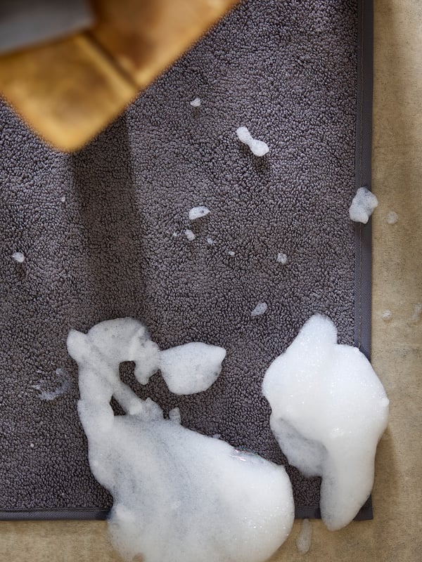 A grey bathroom mat with white soap suds on it. The mat is slightly crumpled and appears soft.
