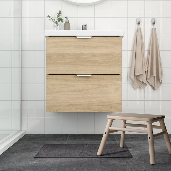 Modern bathroom with wood cabinet, white sink, hanging towels, glass shower, and small black bath mat.