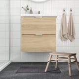 Modern bathroom with wood cabinet, white sink, hanging towels, glass shower, and small black bath mat.