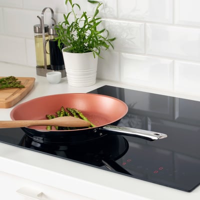 A vibrant red frying pan cooks asparagus on a sleek black induction hob. Wooden utensils, a potted plant, and oils suggest a modern kitchen setup.