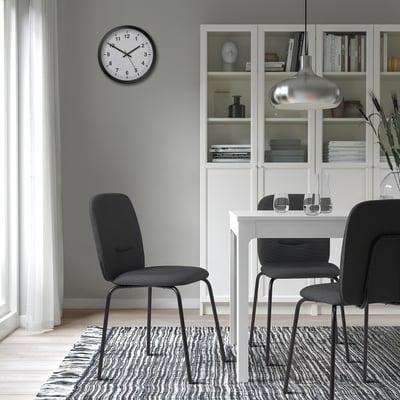 Modern dining area with white table, 3 black PÅBODA chairs, clock, bookshelf, rug, and pendant light.