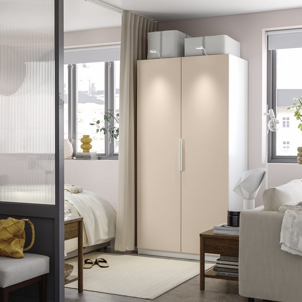 Modern bedroom with beige PAX wardrobe, white bed, gray chair, wooden table, rug, and plants. Wardrobe towers over other items.