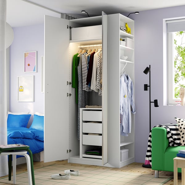 Open wardrobe showing clothes, shelves, and drawers in a bedroom with bed, chair, and window.