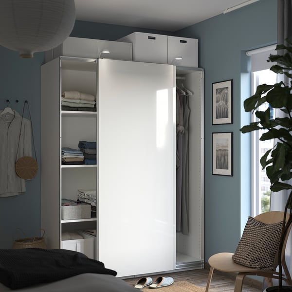 Modern bedroom with PAX white wardrobe, glass door, blue walls, folded clothes, hanging garments, wicker chair, potted plant, and framed photos.