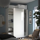 Modern bedroom with PAX white wardrobe, glass door, blue walls, folded clothes, hanging garments, wicker chair, potted plant, and framed photos.