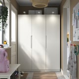 Elegant white PAX wardrobe with integrated lighting, stands tall in room, flanked by narrow shelves and a colorful backpack.
