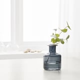 A dark blue glass vase holds green leafy stems near a window, showcasing its use for flowers.
