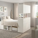 Modern white bedroom with PLATSA bed, shelves, and curtains. Bed centrally placed, surrounded by storage, with a large mirror above.