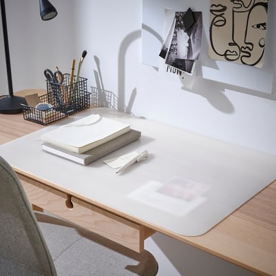 Tidy desk with PLÖJA white pad protecting light wood surface. Notebook, pens, and art prints on walls. Gray chair and lamp nearby.