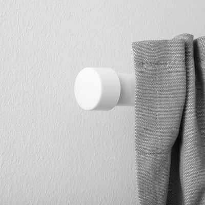 A white cylindrical curtain finial is positioned on a gray curtain rod, attaching the curtain to the wall. The finials design is clean and minimalist.