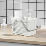 White plastic shower basket with foldable handles, stacked with rolled white towels, placed on light wood countertop beside clear soap dispenser.