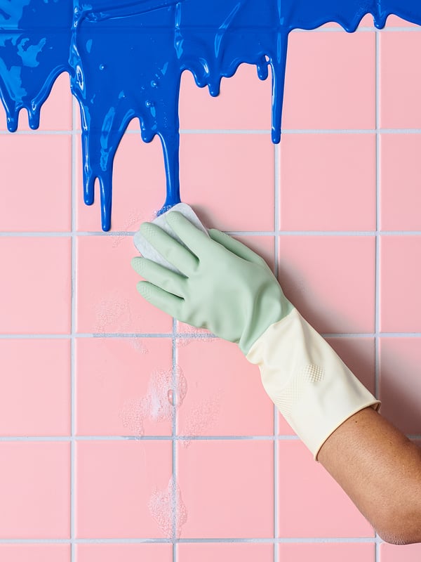 Image shows a gloved hand with green gloves cleaning blue paint off pink tiled wall.