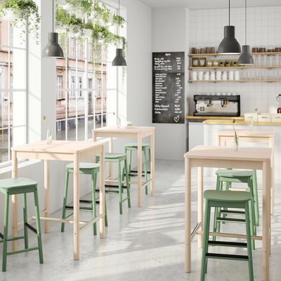 Modern cafe with light wood tables, green stools, hanging lamps, and a chalkboard menu.