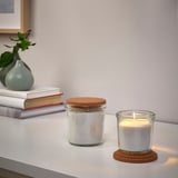Two white candles, one lit, in glass jars with cork lids on a white surface.