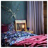 Dark teal bedroom with patterned bedding, striped curtains, hanging blossoms, and a small lamp.