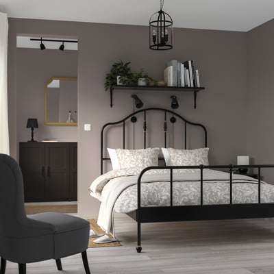 Modern bedroom with black bed, brass details, grey walls, wooden floor, books, plants, grey armchair.