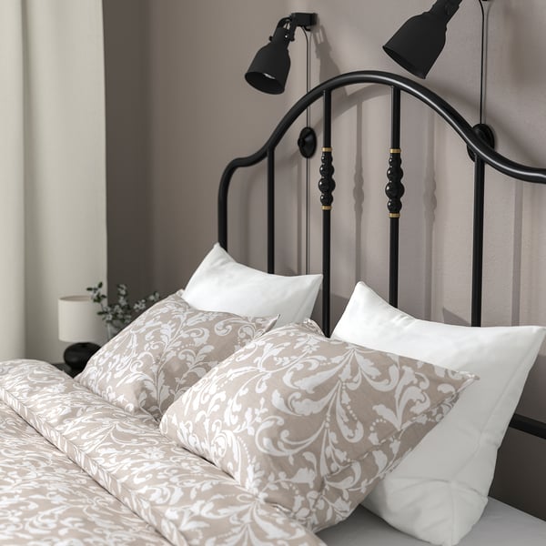 Bed with curved metal headboard, brass details, and wall-mounted lights. Pillows and printed bedsheet are neatly arranged.