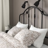Bed with curved metal headboard, brass details, and wall-mounted lights. Pillows and printed bedsheet are neatly arranged.