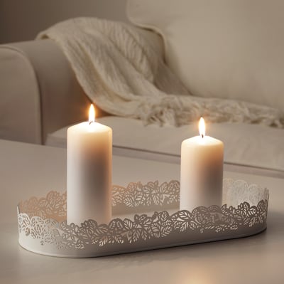 Two lit candles sit in an oval, white, laser-cut metal tray with leaf patterns, adding romantic shadows and ambiance to a light wooden table.