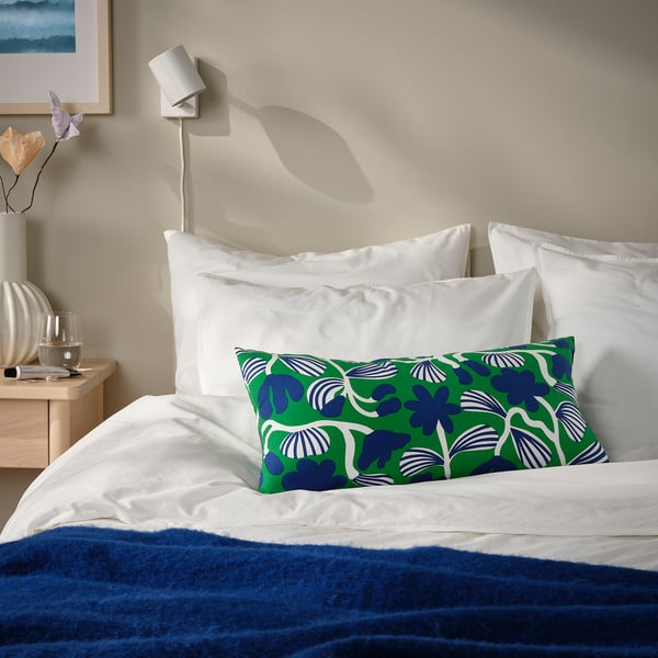 Bed with SANDMOTT green/blue botanical cushion, white linens, blue blanket, wall light, and vase of flowers.